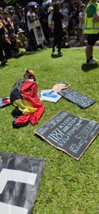 Invasion Day Rally Meanjin Brisbane 2026. Signs lay on the grass before the march. A young person draped in a flag writes on a cutout head of David Crisafulli the text "Our children need just and care, not cruelty and racism."