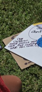 Invasion Day Rally Meanjin Brisbane 2026. Signs lay on the grass before the march. A young person draped in a flag writes on a cutout head of David Crisafulli the text "Our children need just and care, not cruelty and racism."