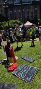 Invasion Day Rally Meanjin Brisbane 2026. Signs lay on the grass before the march. A photographer takes a photo of a young person draped in a flag with a cutout head of David Crisafulli.