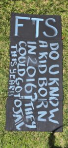 Invasion Day Rally Meanjin Brisbane 2026. Signs lay on the grass before the march. A cardboard sign painted black with blue chalk text says DO U KNOW A KID BORN IN 2016? THEY COULD GO TO JAIL THIS YEAR!! FTS