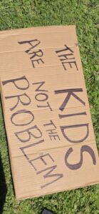 Invasion Day Rally Meanjin Brisbane 2026. Signs lay on the grass before the march. A cardboard sign says THE KIDS ARE NOT THE PROBLEM.