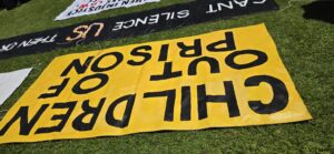 Invasion Day Rally Meanjin Brisbane 2026. Children out of prison sign, black writing on yellow background, on grass before really starts.