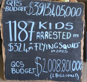 Invasion Day Rally Meanjin Brisbane 2026. Cardboard sign with black paint and blue chalk writing saying QPS Budget $3, 913, 405, 000. 1187 kids arrested by $32.4M "Flying Squad" in 2025. QCS budget $2,008,801,000 ($2 billion)